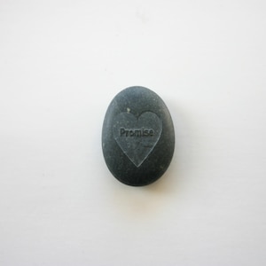 Puede incluir: Una piedra lisa de color gris oscuro con un corazón tallado. La palabra "Promise" está inscrita dentro del corazón.