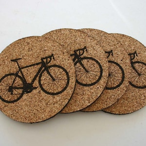 Puede incluir: Conjunto de cuatro posavasos de corcho con una silueta negra de una bicicleta. Cada posavasos tiene un diseño diferente de la bicicleta.