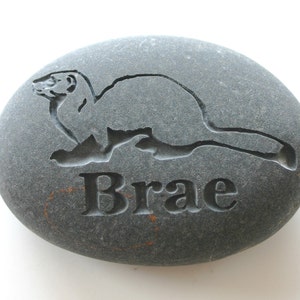 Puede incluir: Una piedra gris con una imagen tallada de una comadreja y el nombre "Brae" grabado debajo.