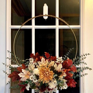 May include: A fall-themed wreath with a gold metal ring and a jute rope top. The wreath features a variety of faux leaves in shades of red, orange, and cream, along with white and green accents. The wreath is hanging on a white window.