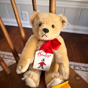 May include: A plush, golden-brown teddy bear with a red ribbon around its neck. The bear has black eyes and a black stitched nose. A tag from Hamleys, featuring a cartoon figure, is attached. The bear is seated on a wooden chair.