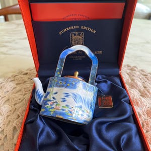 Puede incluir: Una tetera de porcelana azul y blanca con asa y tapa doradas. La tetera está decorada con un diseño floral y se encuentra en una caja roja con forro de satén azul. La caja tiene una etiqueta que dice "Edición numerada".