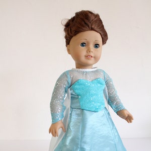 May include: A doll wearing a light blue gown with a white cape and a sparkly bodice. The doll has brown hair and blue eyes.