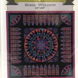 May include: A package featuring a "Rose Window" quilt pattern, measuring 68" x 68". The design includes a central circular motif and surrounding rectangular panels in shades of blue, red, and purple. The package is labeled "Mariner's Compass".