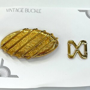 May include: Gold-toned vintage buckle with a textured oval shape and a smaller, rectangular buckle. The card reads "Vintage Buckle".