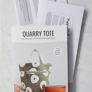 May include: A sewing pattern for a "QUARRY TOTE" with a flat bottom design and a bonus zipped pouch. The tote is grey with white circular patterns. The image includes additional pattern sheets and the text "noodlehead".
