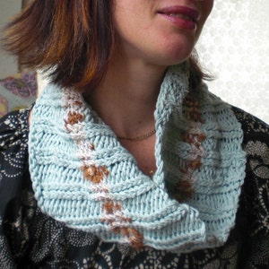 May include: A light blue crocheted cowl with brown and white accents. The cowl is folded over and worn around the neck.