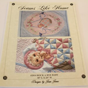 May include: A quilting pattern for a baby quilt titled "Rock A Bye Baby" by Joan Jones. The pattern includes a pink and blue baby quilt with a baby in a crib and a close-up of the baby's face. The quilt measures 96.5 cm long by 73.7 cm wide.