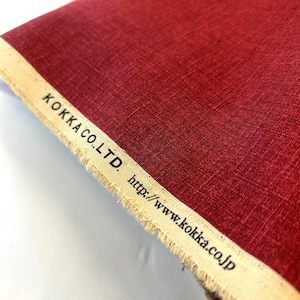 May include: A deep red fabric with a visible woven texture and a frayed, off-white selvage edge. The selvage is printed with the black text KOKKA CO.,LTD. and the website address http://www.kokka.co.jp along the length of the material.