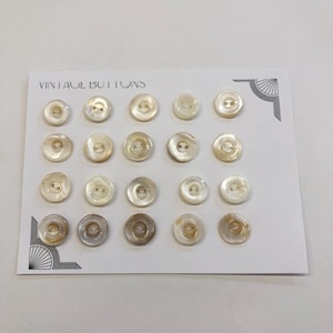 May include: A collection of vintage buttons arranged on a white card. The buttons are round with a pearlescent sheen, varying in shades of white, cream, and gray. The card is labeled "VINTAGE BUTTONS" and features decorative corner designs.