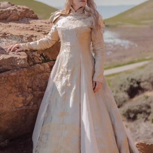 May include: A long, cream-colored gown with intricate gold embroidery. The dress features long sleeves, a high neckline, and a full skirt. A silver crown and pearl necklace complete the regal look. The setting is outdoors, with a rocky landscape.