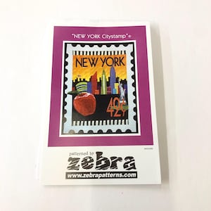 May include: A pink and white paper pattern for a quilt featuring a stylised image of the New York City skyline with a red apple and the text "NEW YORK" and "42c". The pattern is titled "NEW YORK Citystamp" and is patterned by zebra.