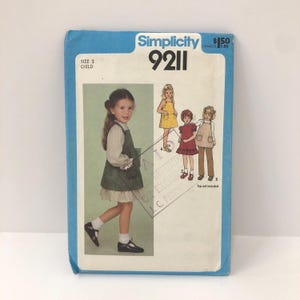 Può includere: Modello di cucito Simplicity 9211 per un vestito da bambina. La copertina presenta una foto di una bambina con un vestito verde sopra una camicetta bianca e disegni di altri tre modelli di vestiti. Taglia 3.