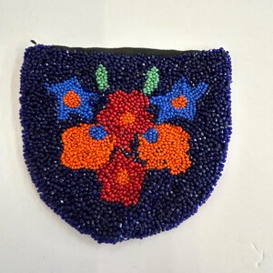 May include: A small, semi-circular pouch covered in dark blue beads. The front features a floral pattern created with orange, red, and light blue beads, accented by two small green leaf shapes at the top. A black zipper is visible along the top edge.