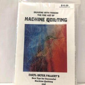 May include: A book titled "Drawing with Thread: The Fine Art of Machine Quilting" is sealed in a clear plastic bag. The cover features a hummingbird design with blue, red, and orange hues. The book also includes "Best Tips for Successful Machine Quilting" and 12 practice patterns.