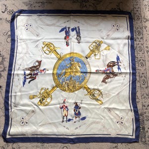 May include: Square silk scarf with a white background, navy blue border, and a central design featuring a gold emblem with a knight on horseback. The scarf has gold swords and illustrations of soldiers and horses.