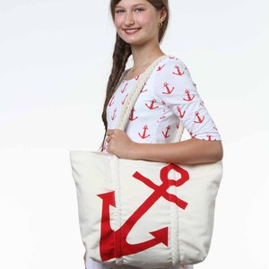 Op de afbeelding: Een witte canvas tote bag met een groot rood ankerontwerp. De tas heeft touwhandvatten en wordt vastgehouden door een persoon die een wit shirt draagt met een rood ankerpatroon.