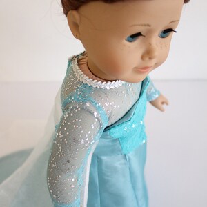 May include: A doll wearing a light blue dress with silver glitter and a white lace trim. The dress has long sleeves and a flowing skirt.