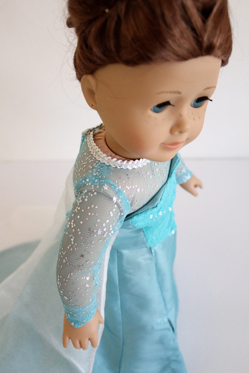 May include: A doll wearing a light blue dress with silver glitter and a white lace trim. The dress has long sleeves and a flowing skirt.