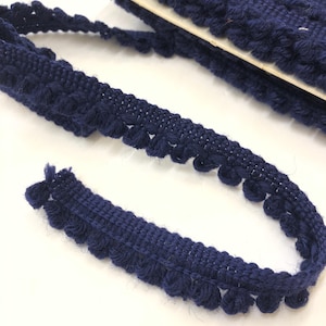 May include: Close-up of navy blue pom-pom trim. The trim is made of a woven fabric with a row of small, evenly spaced pom-poms along one edge. The trim is partially rolled up, with a section unraveled, showing the texture and detail of the material.