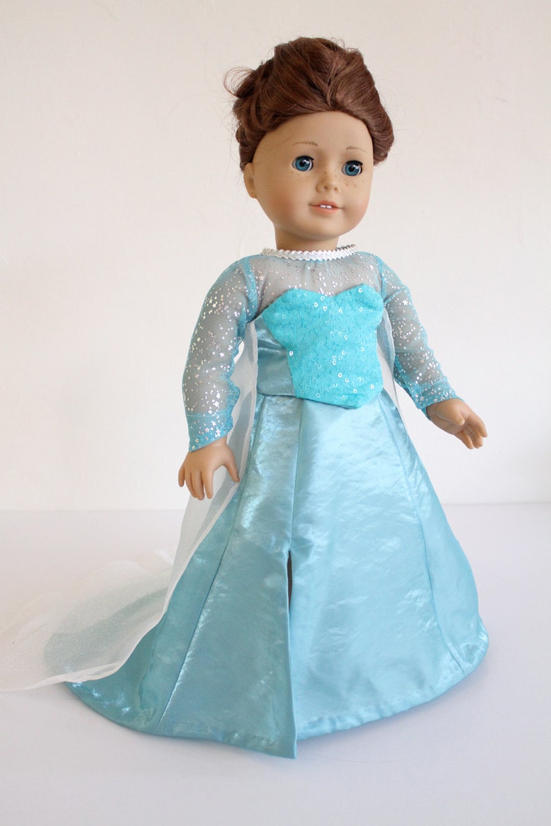 May include: A doll wearing a light blue gown with a white cape and a sparkly bodice. The doll has brown hair and blue eyes.