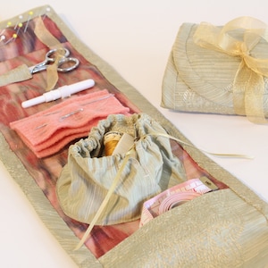 Op de afbeelding: Een naaiset van beige stof met een roze voering van stof en een verscheidenheid aan naai-gereedschap, waaronder naalden, garen, een schaar en een meetlint. De set is opgerold en vastgebonden met een gouden lint.
