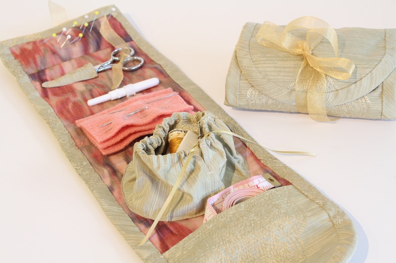 Op de afbeelding: Een naaiset van beige stof met een roze voering van stof en een verscheidenheid aan naai-gereedschap, waaronder naalden, garen, een schaar en een meetlint. De set is opgerold en vastgebonden met een gouden lint.