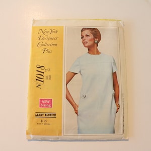 May include: A vintage sewing pattern for a dress, featuring a woman wearing a light blue dress with short sleeves. The pattern is from the New York Designers Collection Plus and is size 12 with a bust measurement of 34 inches. The pattern is labeled N1018 and is by Larry Aldrich.