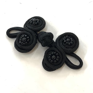 May include: Black frog closure with a spiral design. The closure features a central knot and two decorative spiral elements with beaded centers. The frog closure is made of a woven material.