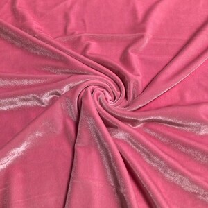 Puede incluir: Primer plano de una tela de terciopelo rosa con un brillo suave y lustroso. La tela está reunida en espiral, creando pliegues y sombras que resaltan su textura. El color es un rosa medio vibrante.