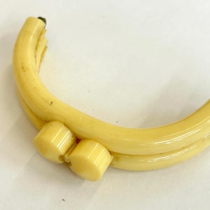 May include: A curved, pale yellow plastic purse frame featuring a dual-hinged design with two circular knobs at the center for opening and closing. The frame has a smooth, glossy finish and a semi-circular shape, designed for attaching to a bag.