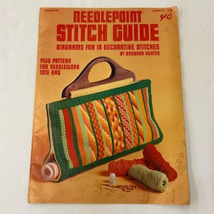 May include: Vintage "Needlepoint Stitch Guide" book with diagrams for decorative stitches and a tote bag pattern. The cover is orange with red text. A needlepoint tote bag with a wooden handle is shown, along with yarn, scissors, and a thimble.
