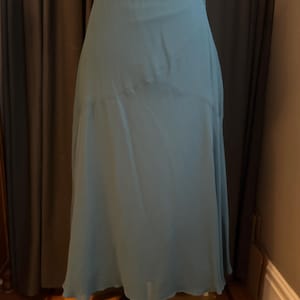 Puede incluir: Una falda fluida de color verde azulado, de cintura alta. La falda está hecha de una tela ligera que se drapea con gracia. La falda se muestra en un maniquí, mostrando su forma y diseño. El dobladillo de la falda llega a la mitad de la pantorrilla.