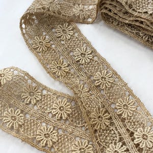 May include: Close-up of a roll of gold-colored lace trim. The trim features a floral design with detailed openwork. The lace is likely made of a delicate material, perfect for sewing and craft projects.