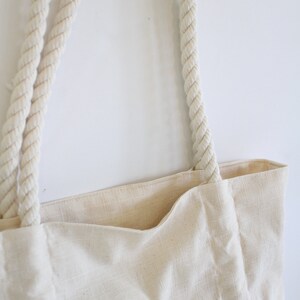 May include: A white canvas tote bag with thick, white rope handles. The bag is made of a natural fabric with a subtle texture.