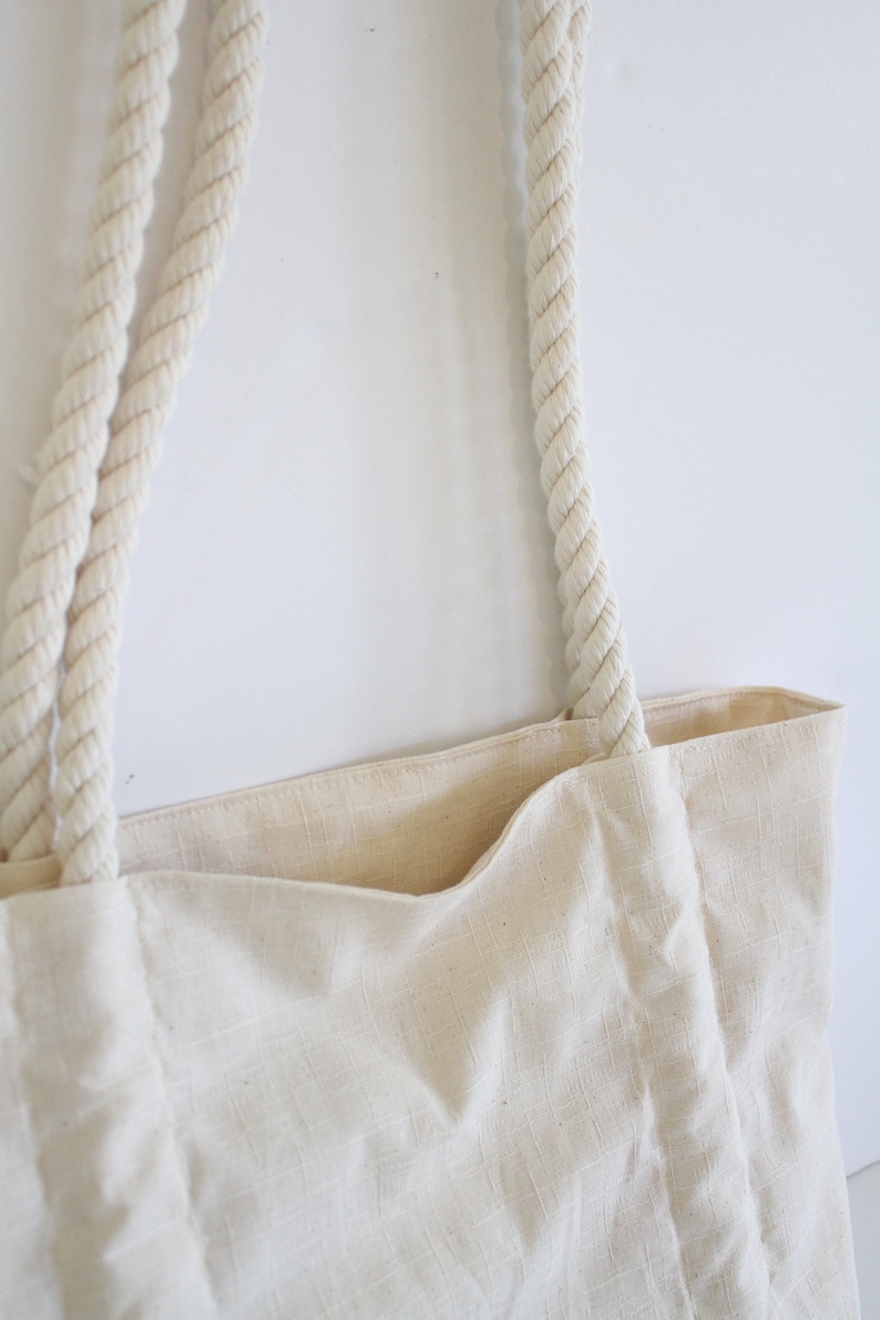 May include: A white canvas tote bag with thick, white rope handles. The bag is made of a natural fabric with a subtle texture.