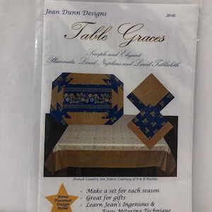 May include: A package for "Table Graces" by Jean Dunn Designs. The package features images of placemats, napkins, and a tablecloth with a blue and gold floral design. The text on the package includes "Simple and Elegant" and "Make It Easy Sewing & Crafts."