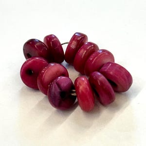 May include: A collection of eleven circular, disc-shaped beads in a vibrant magenta-pink hue. Each bead features a central hole and a smooth, polished surface, arranged in a loose, slightly overlapping cluster on a plain white surface.