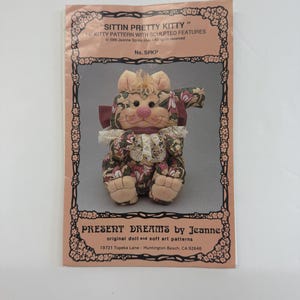 May include: A pattern for a 14 inch sitting kitty doll with sculpted features. The pattern is titled "Sittin Pretty Kitty" and is by Present Dreams by Jeanne. The pattern includes a picture of the finished doll, which is a green, pink, and white cat with a pink bow.