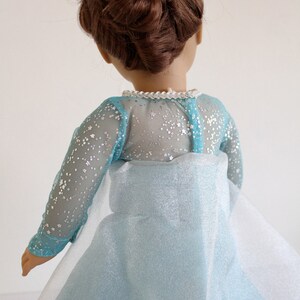 May include: A doll wearing a light blue dress with a sparkly tulle overlay and a silver glitter bodice. The dress has long sleeves and a high neckline. The doll has brown hair styled in an updo.