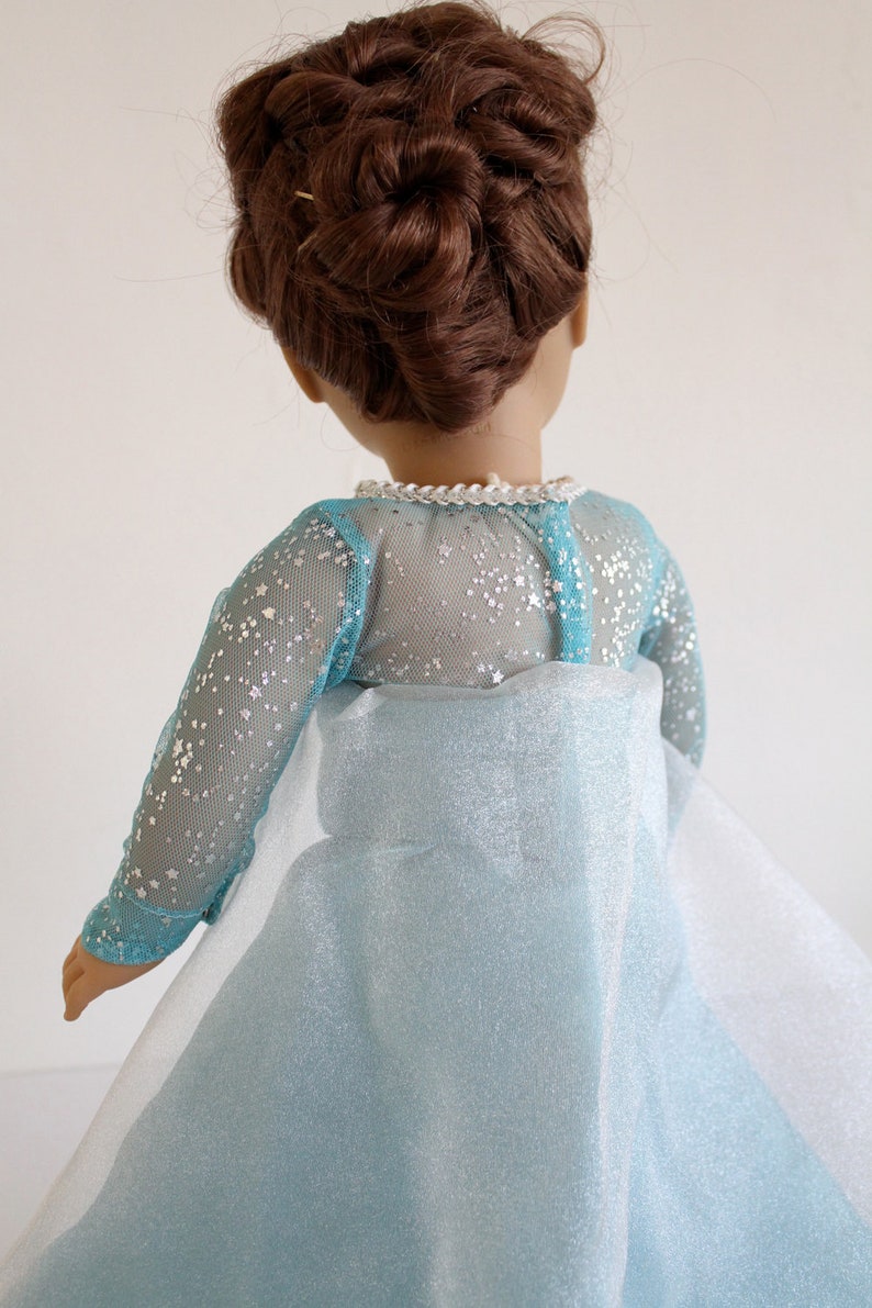 May include: A doll wearing a light blue dress with a sparkly tulle overlay and a silver glitter bodice. The dress has long sleeves and a high neckline. The doll has brown hair styled in an updo.