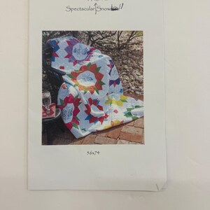 May include: A close-up of a colorful quilt with a starburst pattern. The quilt is laid out on a brick patio and is surrounded by green plants. The text "Frame for Spectacular Snowball!" is written on the image. The text "56x74" is also written on the image.