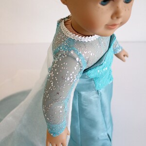 May include: A doll wearing a light blue dress with a white lace trim and silver glitter. The dress has long sleeves and a flowing skirt.