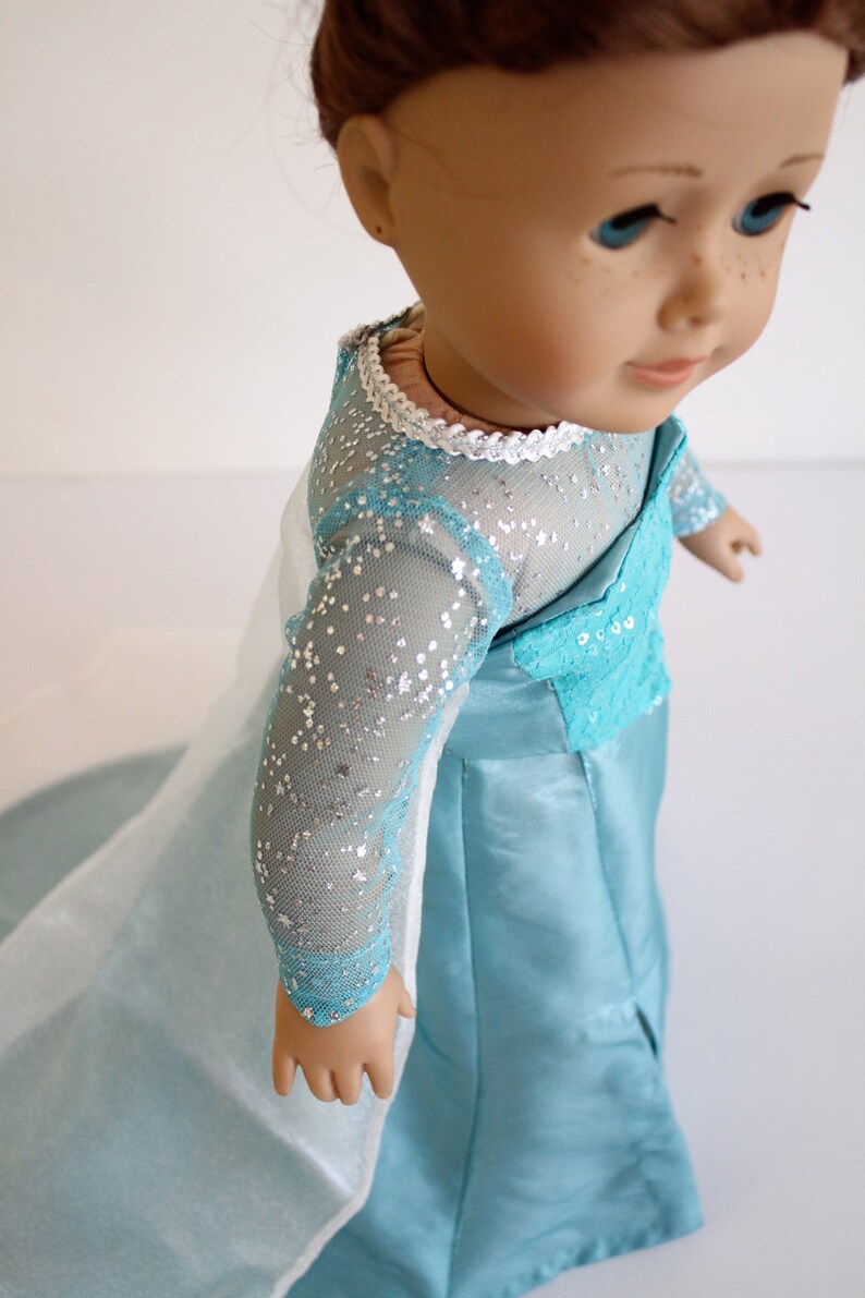 May include: A doll wearing a light blue dress with a white lace trim and silver glitter. The dress has long sleeves and a flowing skirt.