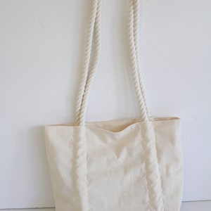May include: A white canvas tote bag with thick rope handles. The bag has a simple design and is made of a natural fabric.
