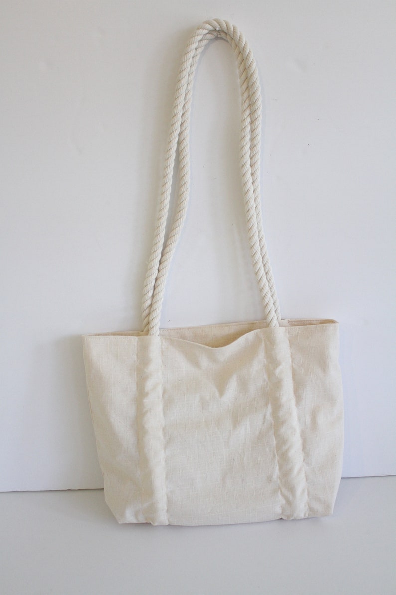 May include: A white canvas tote bag with thick rope handles. The bag has a simple design and is made of a natural fabric.
