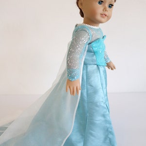 May include: A doll wearing a light blue dress with a long, flowing train and a sparkly bodice. The dress has a white tulle overlay. The doll has brown hair and blue eyes.