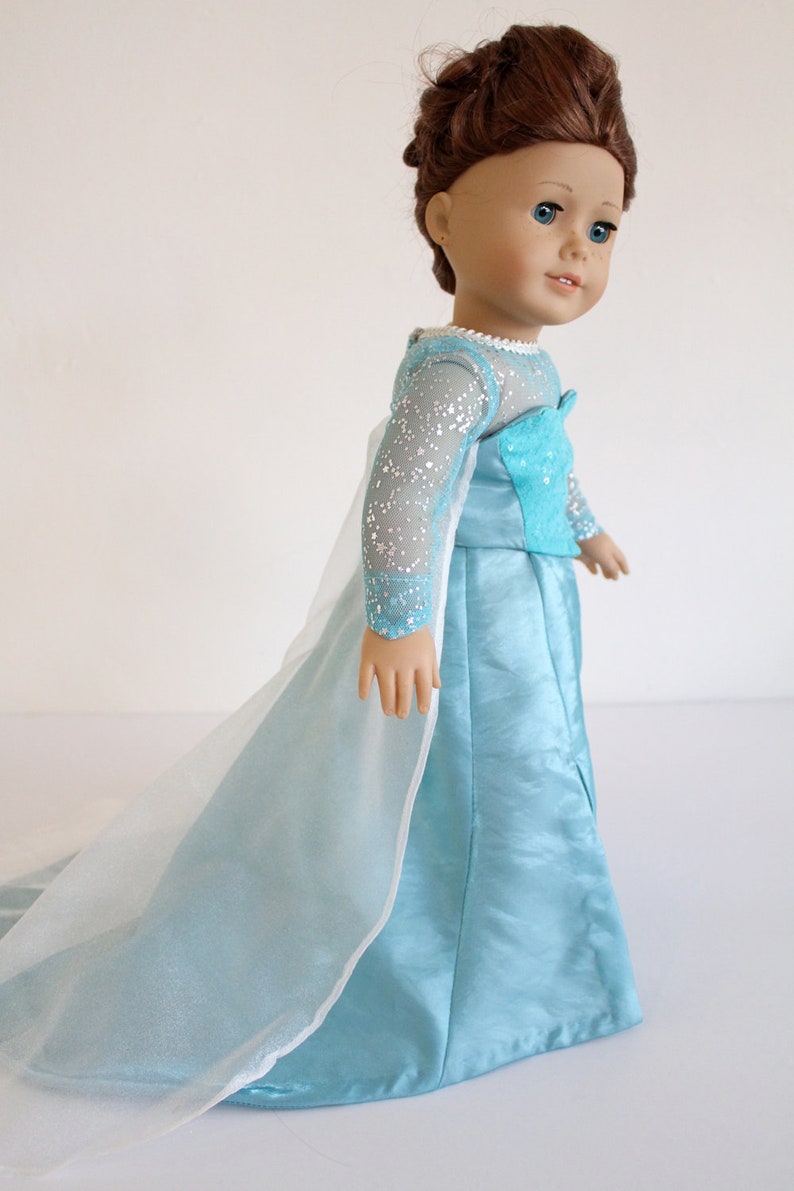 May include: A doll wearing a light blue dress with a long, flowing train and a sparkly bodice. The dress has a white tulle overlay. The doll has brown hair and blue eyes.