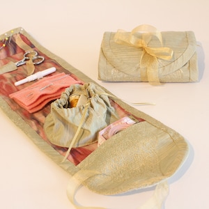 Op de afbeelding: Een beige stoffen naaiset met een roze en wit gepatroneerd interieur. De set is open en bevat verschillende naai benodigdheden, waaronder naalden, draad, een schaar en een vingerhoed. De set is vastgebonden met een gouden lint.