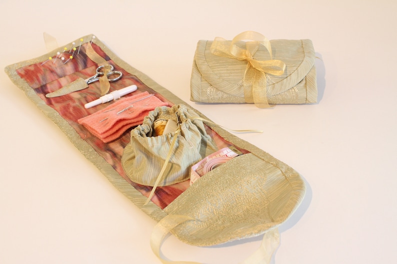 Op de afbeelding: Een beige stoffen naaiset met een roze en wit gepatroneerd interieur. De set is open en bevat verschillende naai benodigdheden, waaronder naalden, draad, een schaar en een vingerhoed. De set is vastgebonden met een gouden lint.